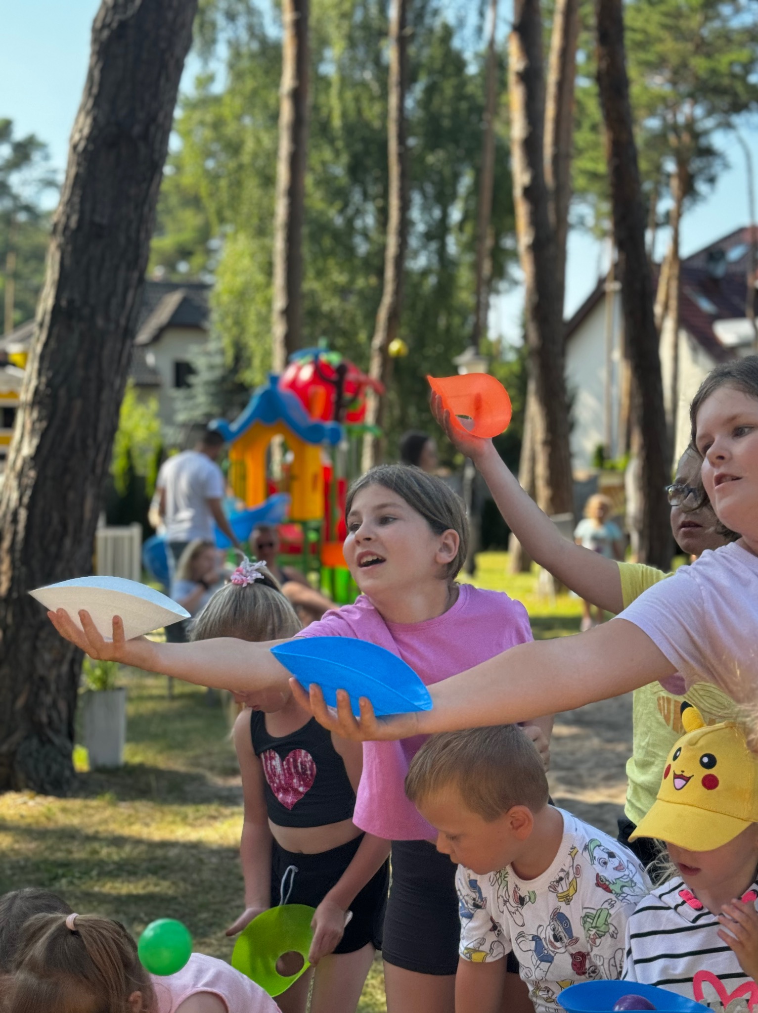 Grupa dzieci w różnym wieku bawi się w parku, trzymając kolorowe, plastikowe talerze w dłoniach podczas zabawy z piłeczkami.