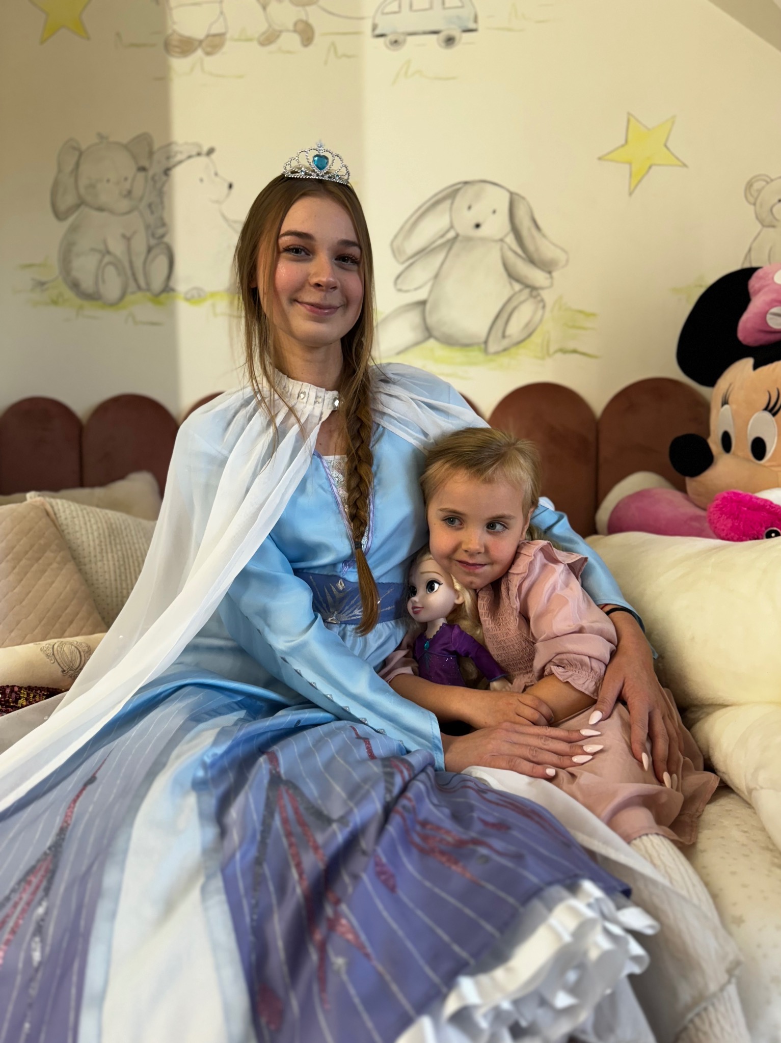Animator w stroju Elsy z Krainy Lodu obejmuje dziewczynkę trzymającą lalkę Elsy na tle ściany z dziecięcymi rysunkami.