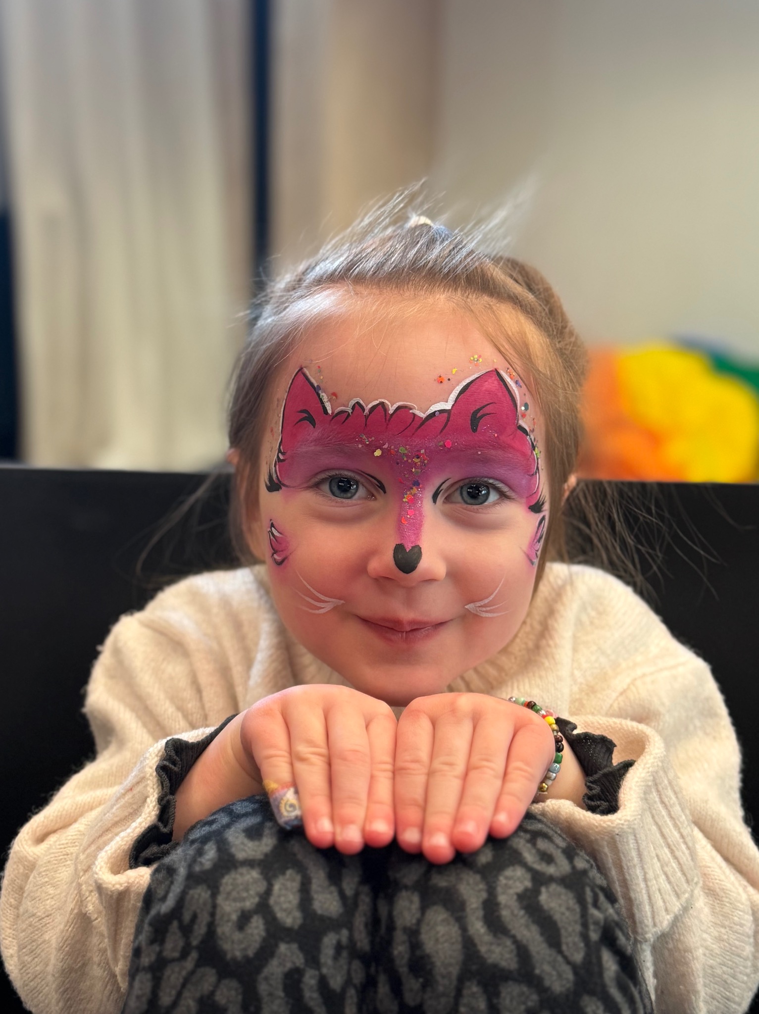 Uśmiechnięta dziewczynka z pomalowaną twarzą w motywy różowego liska, brokat i białe wąsy. Dziecko opiera ręce na kolanach, w tle widoczny kolorowy element.