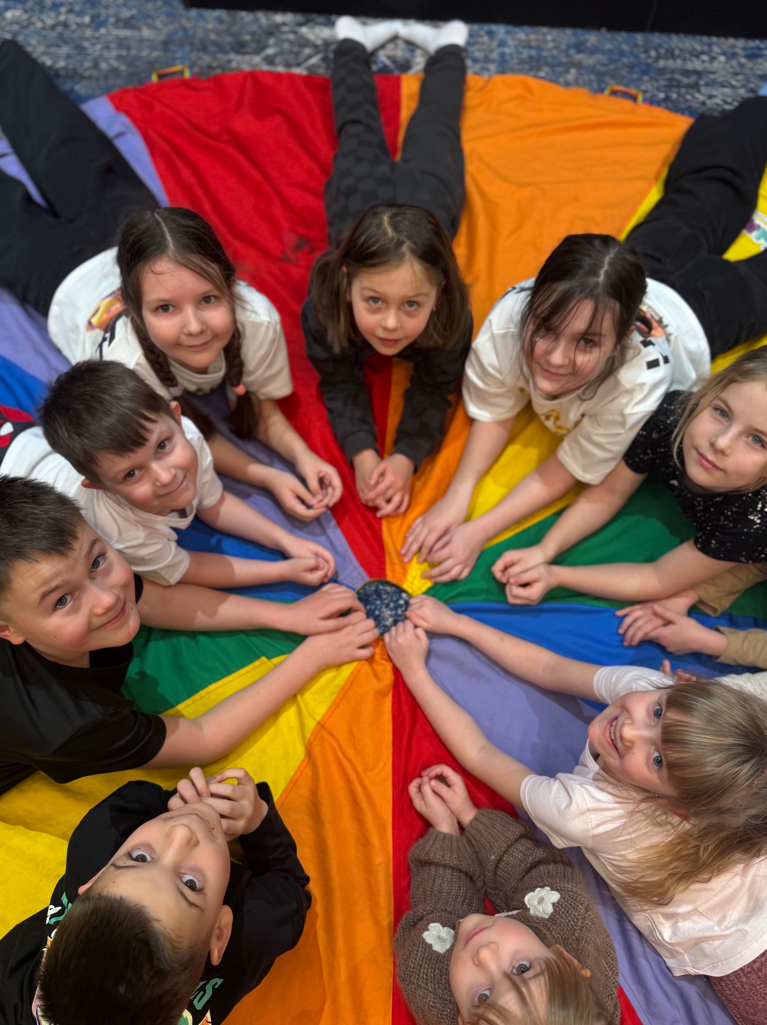 Grupka dzieci leżących na kolorowej chuście animacyjnej, tworzących krąg. Uśmiechnięte twarze skierowane w górę, dłonie w centrum koła. Zabawa integracyjna.