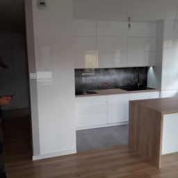 Aneta-meble - Montaż białej kuchni z wyspą: widoczny monter w kombinezonie sprawdzający coś na smartfonie na tle nowoczesnej zabudowy kuchennej z ciemnym panelem nad blatem.