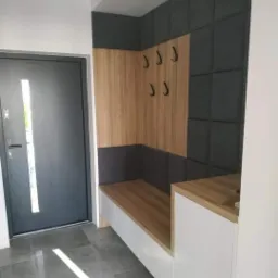 Wnętrze przedpokoju z zabudową: drewnopodobna płyta na ścianie z wieszakami, szare panele tapicerowane, białe szafki z siedziskiem z płyty drewnopodobnej, szara podłoga z płytek, ciemne drzwi...