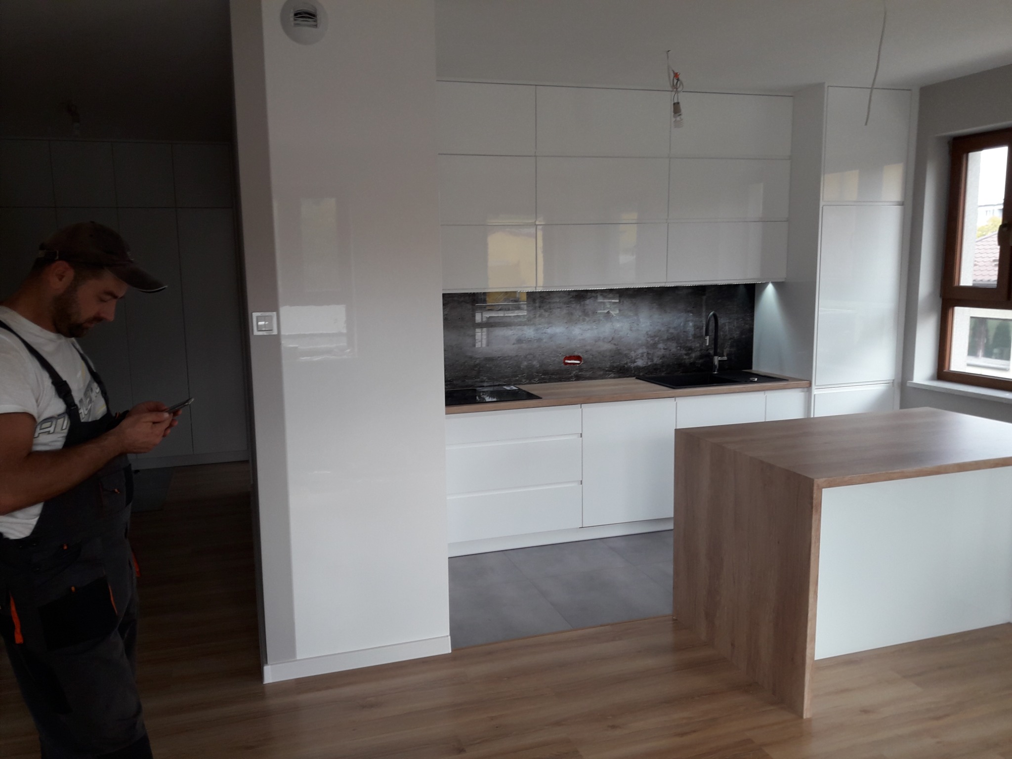 Montaż białej kuchni z wyspą: widoczny monter w kombinezonie sprawdzający coś na smartfonie na tle nowoczesnej zabudowy kuchennej z ciemnym panelem nad blatem.