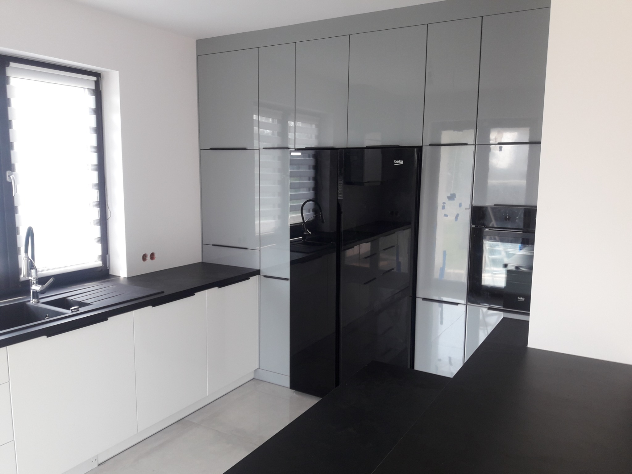 Nowoczesna kuchnia z szarymi szafkami górnymi, białymi dolnymi, czarnym blatem i czarną lodówką, minimalistyczny design.