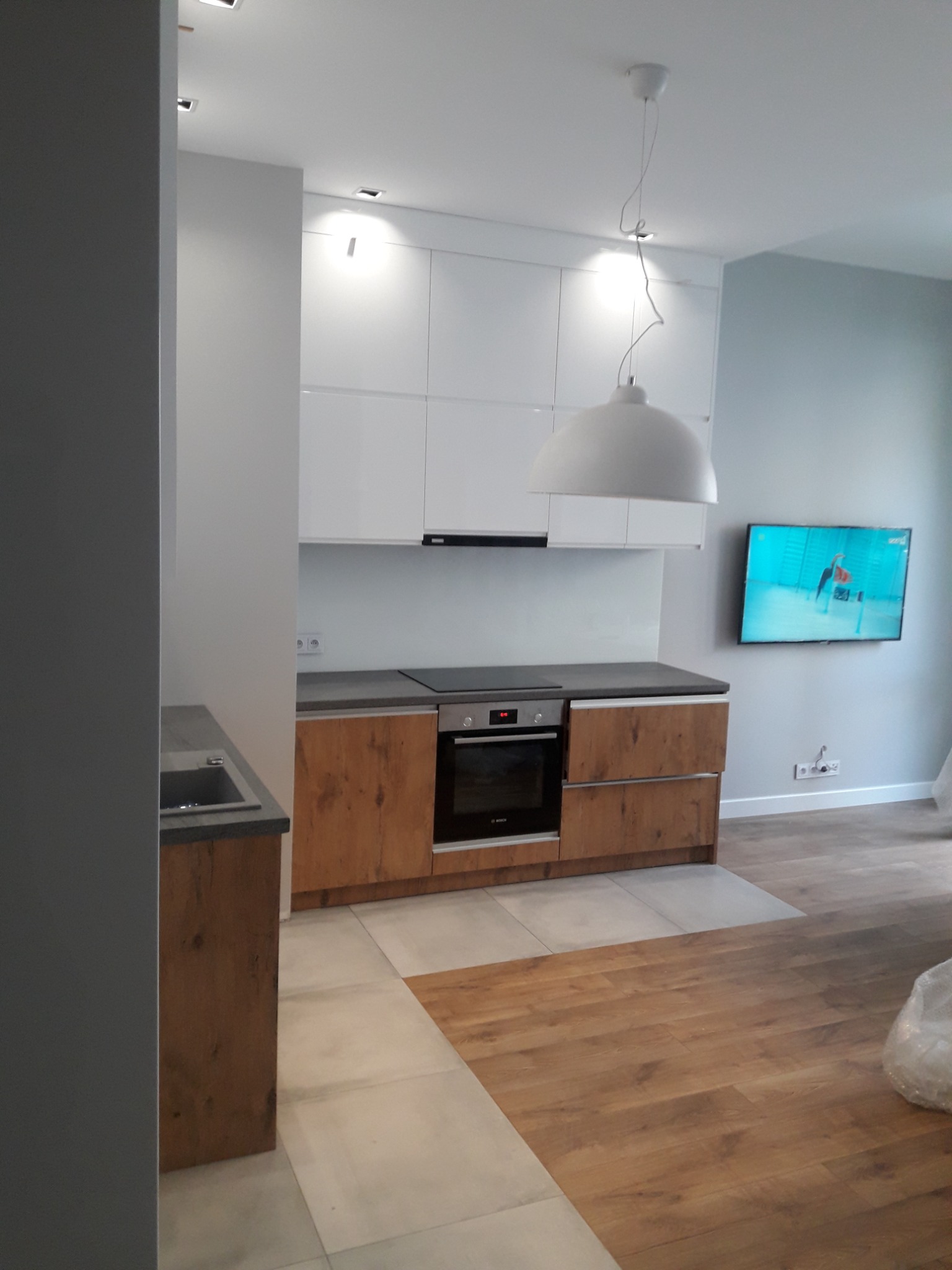 Nowoczesna kuchnia z białymi szafkami wiszącymi, drewnopodobnymi frontami dolnych szafek i szarym blatem, widziana z perspektywy wejścia do pomieszczenia, z widocznym fragmentem podłogi z paneli.