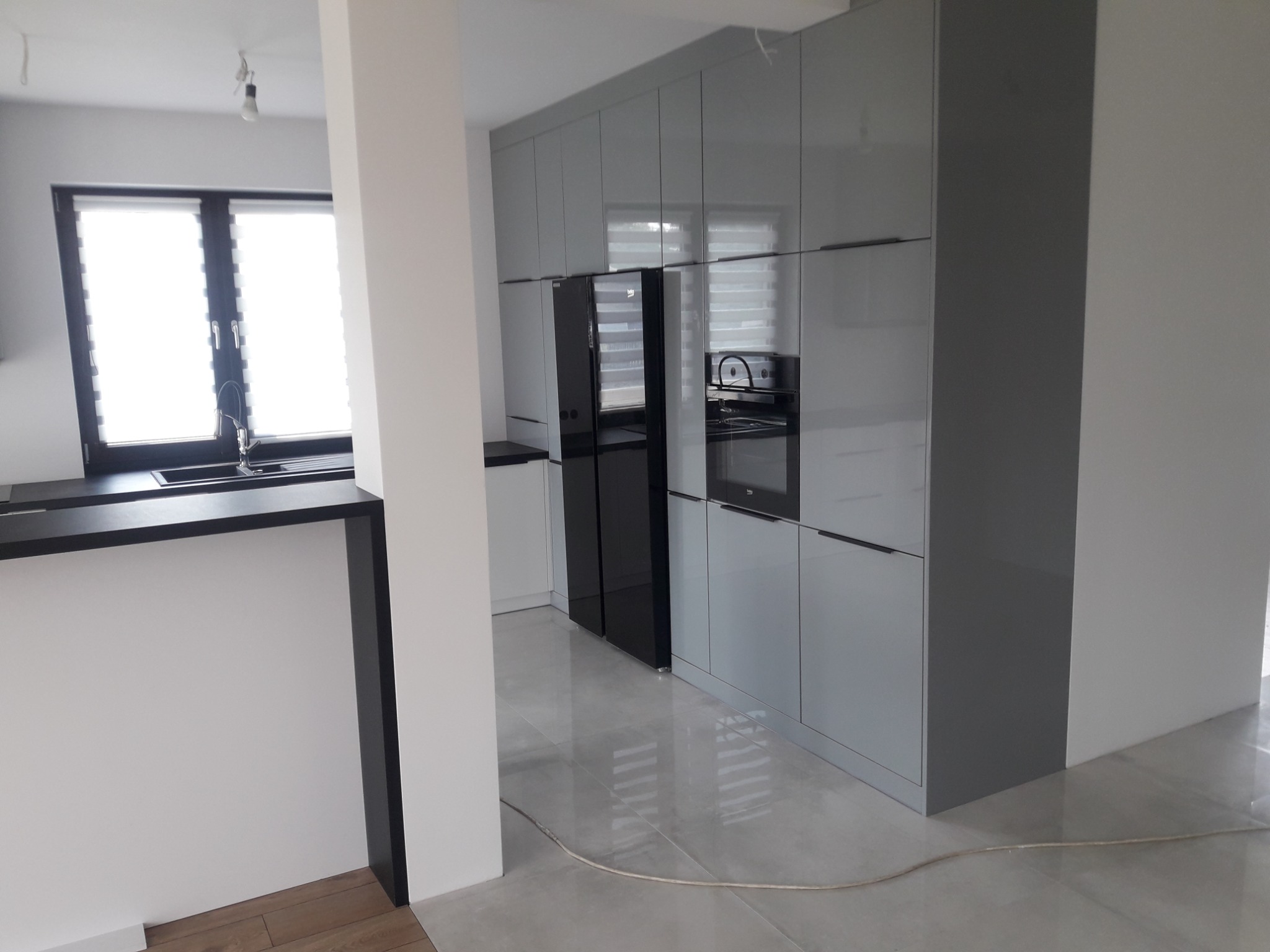 Nowoczesna kuchnia w odcieniach szarości z połyskującymi frontami szafek, czarnym sprzętem AGD i widocznym fragmentem okna z żaluzjami.