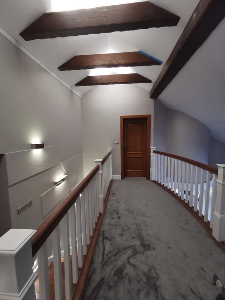 Korytarz na poddaszu po remoncie z szarą wykładziną, białymi balustradami i drewnianymi belkami stropowymi, oświetlony kinkietami.