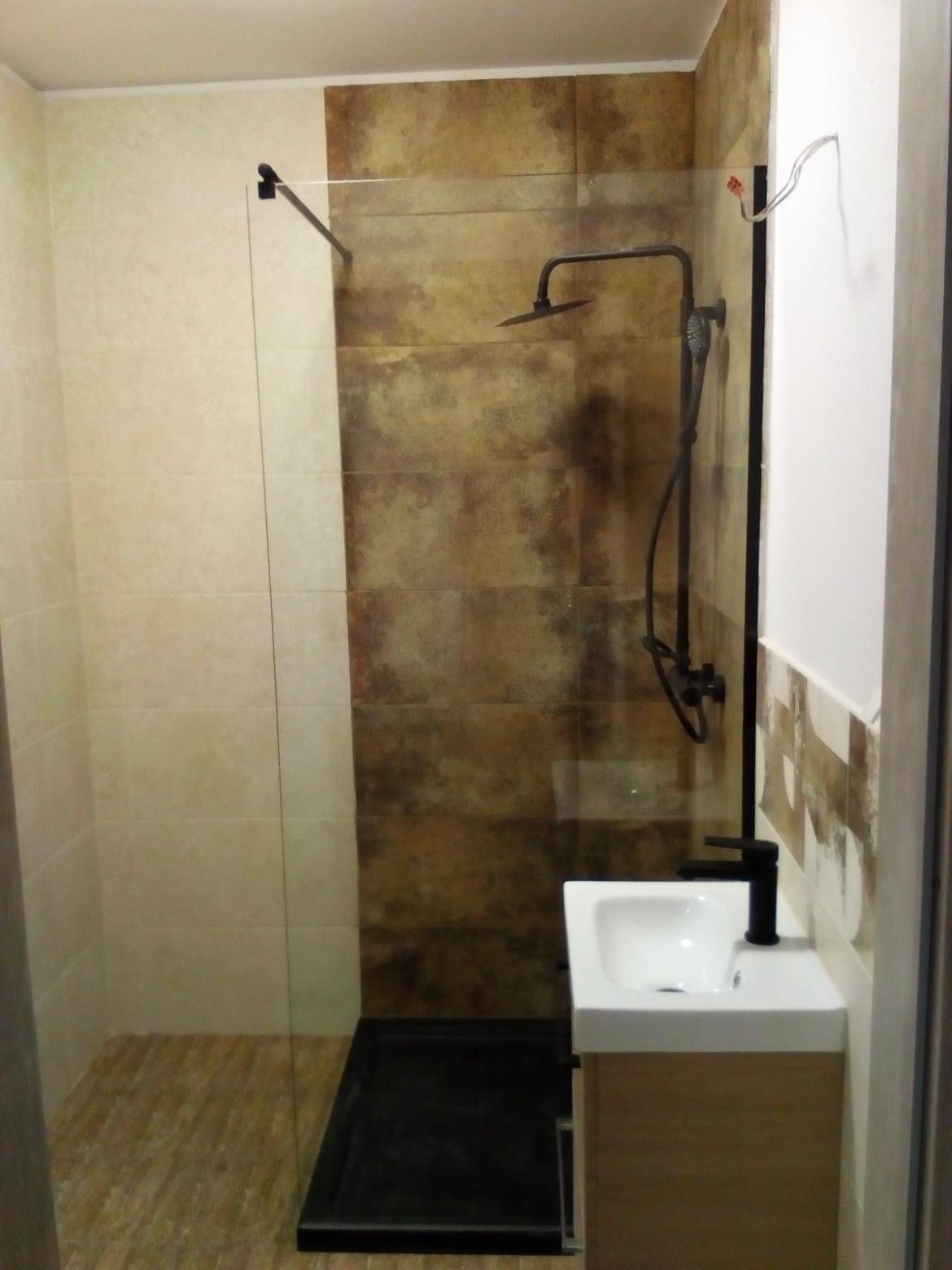 Wykończona łazienka z kabiną prysznicową typu walk-in, ciemną armaturą i umywalką nablatową na szafce z drewnopodobnym frontem. Widoczne płytki w odcieniach beżu i brązu oraz wystające przewody...