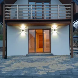 Nowoczesny dom jednorodzinny z drewnianymi elementami wykończeniowymi, oświetleniem elewacyjnym i brukowaną ścieżką prowadzącą do wejścia.