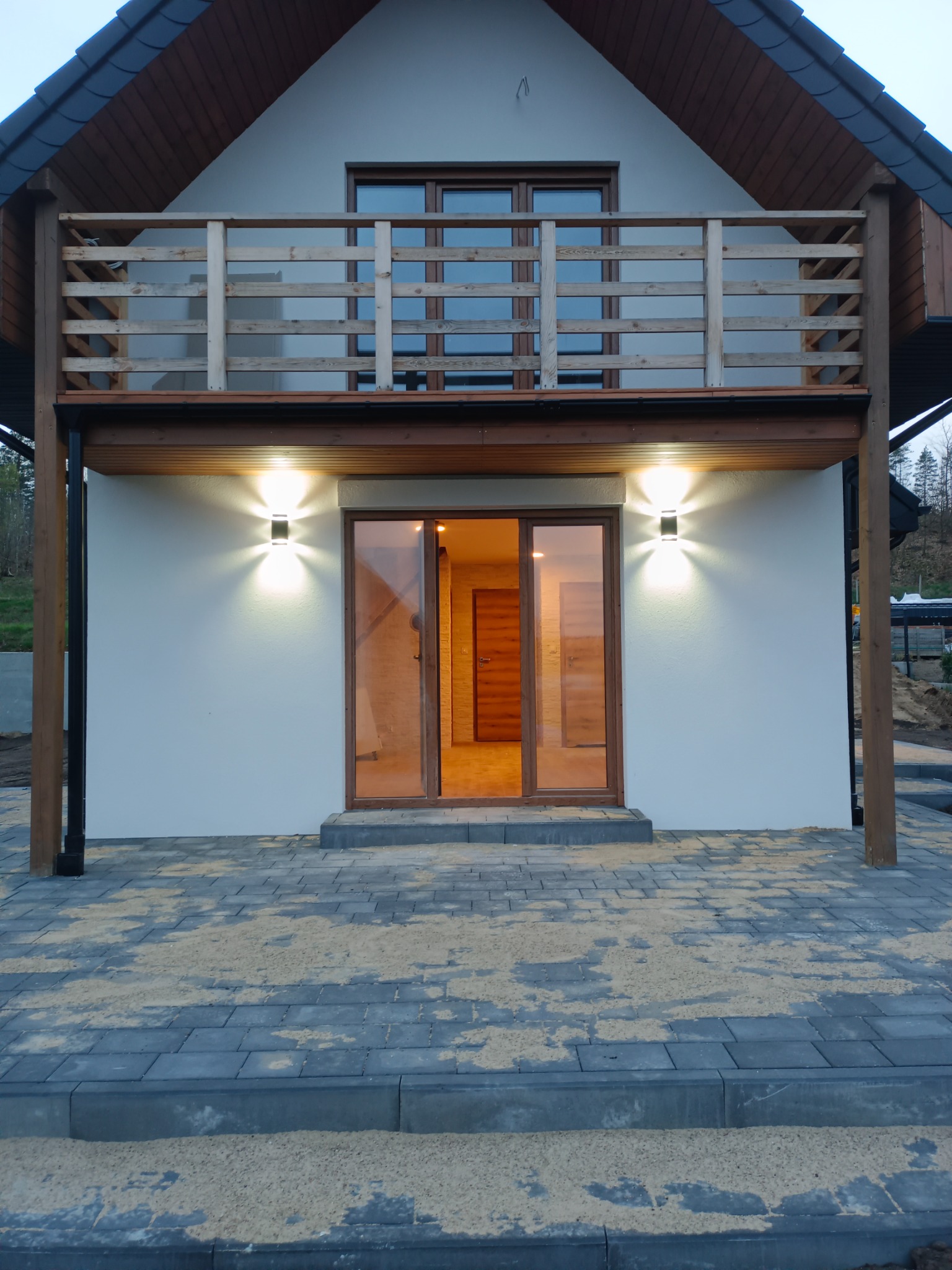 Nowoczesny dom jednorodzinny z drewnianymi elementami wykończeniowymi, oświetleniem elewacyjnym i brukowaną ścieżką prowadzącą do wejścia.