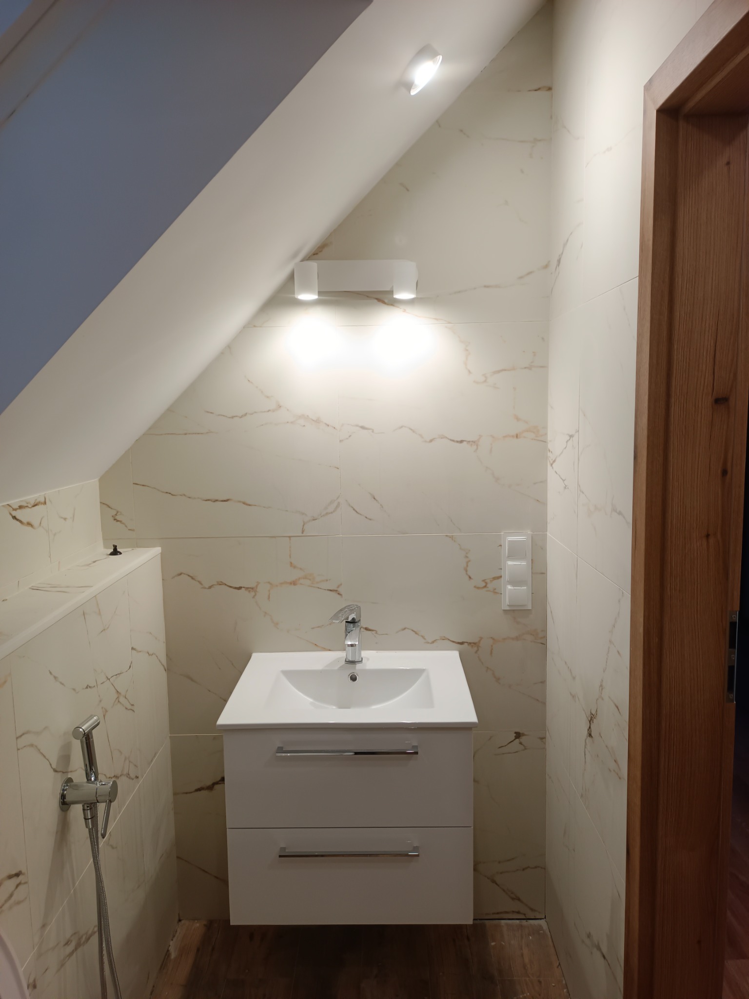 Nowoczesna łazienka na poddaszu z umywalką nablatową na szafce z dwiema szufladami, bidetką i marmurkowymi płytkami oraz oświetleniem punktowym.
