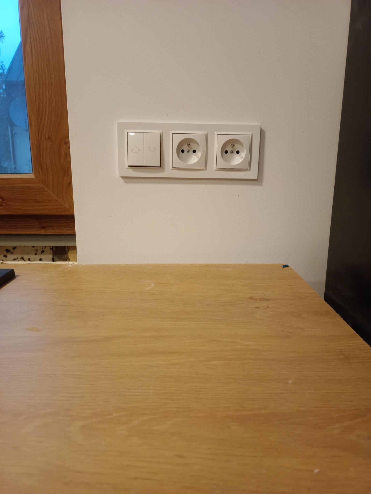 Biały panel ścienny z dwoma gniazdkami elektrycznymi i podwójnym włącznikiem światła, zamontowany nad drewnianym blatem.