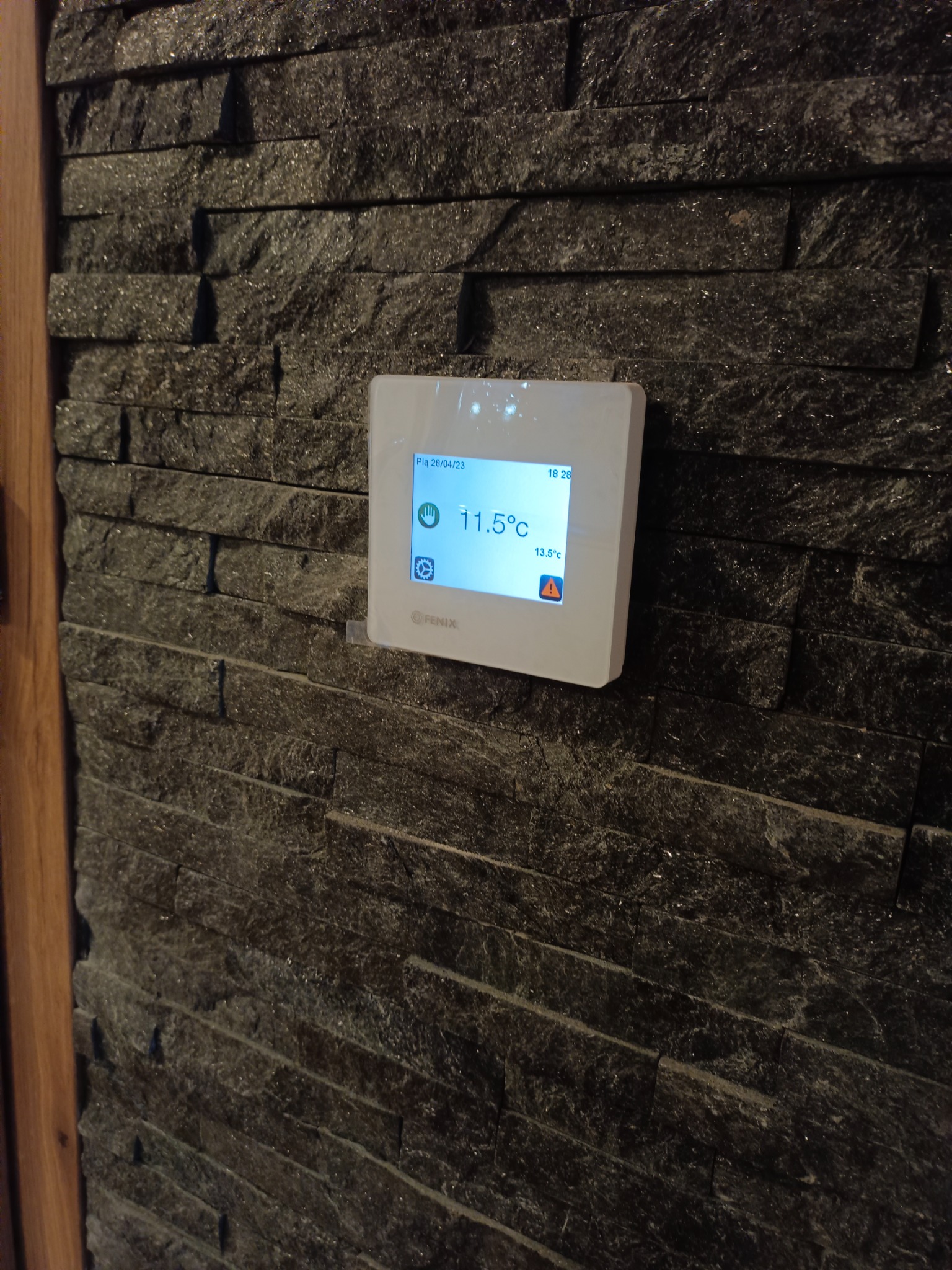 Biały panel sterowania ogrzewaniem podłogowym marki Fenix, zamontowany na dekoracyjnej ścianie z czarnego kamienia w Sławnie, wyświetlający temperaturę 11.5°C.