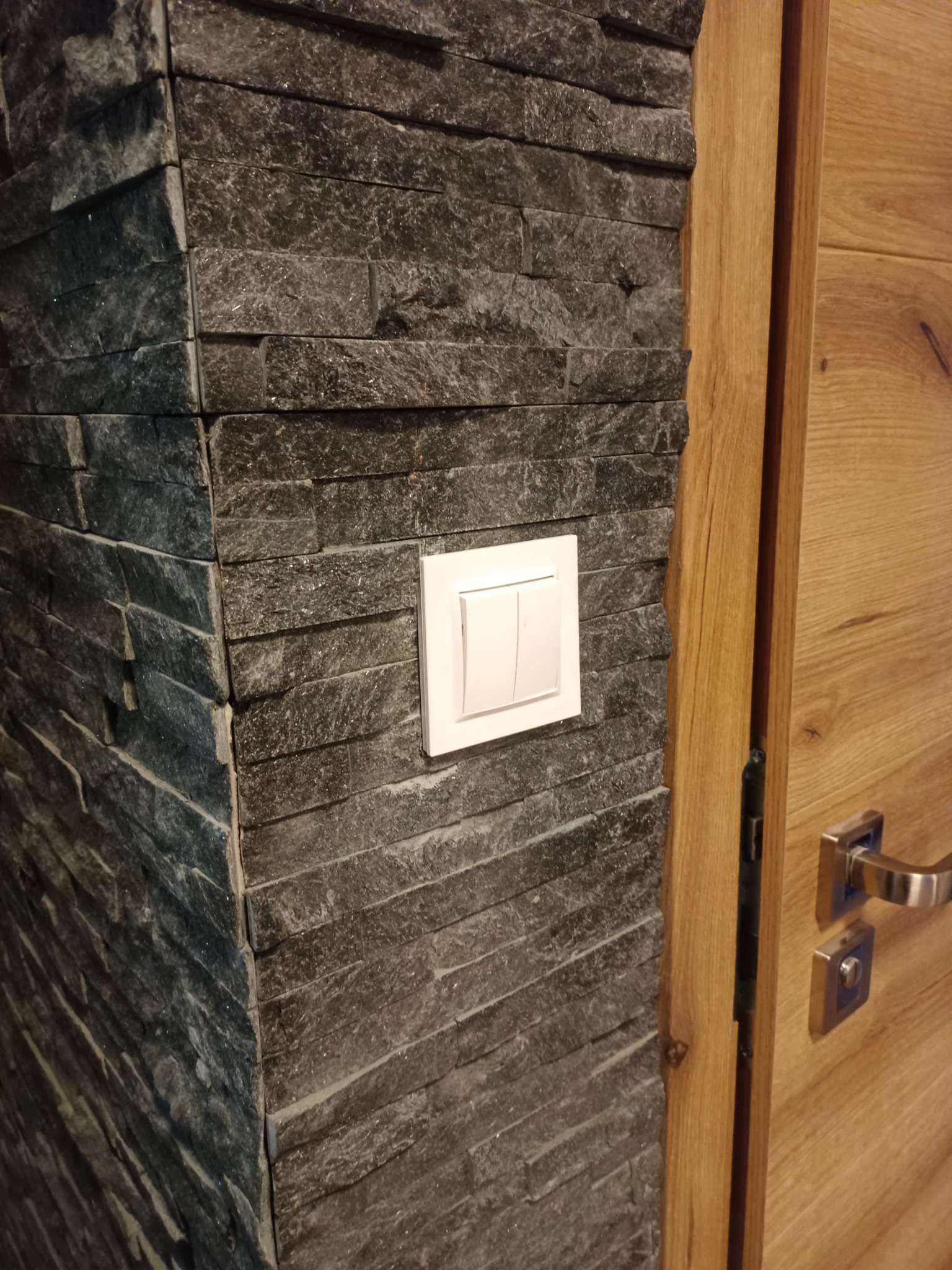 Ściana wykończona szarą, łupaną płytką kamienną z zamontowanym podwójnym, białym włącznikiem światła, przylegająca do drewnianej ościeżnicy i częściowo widocznych drzwi z klamką.