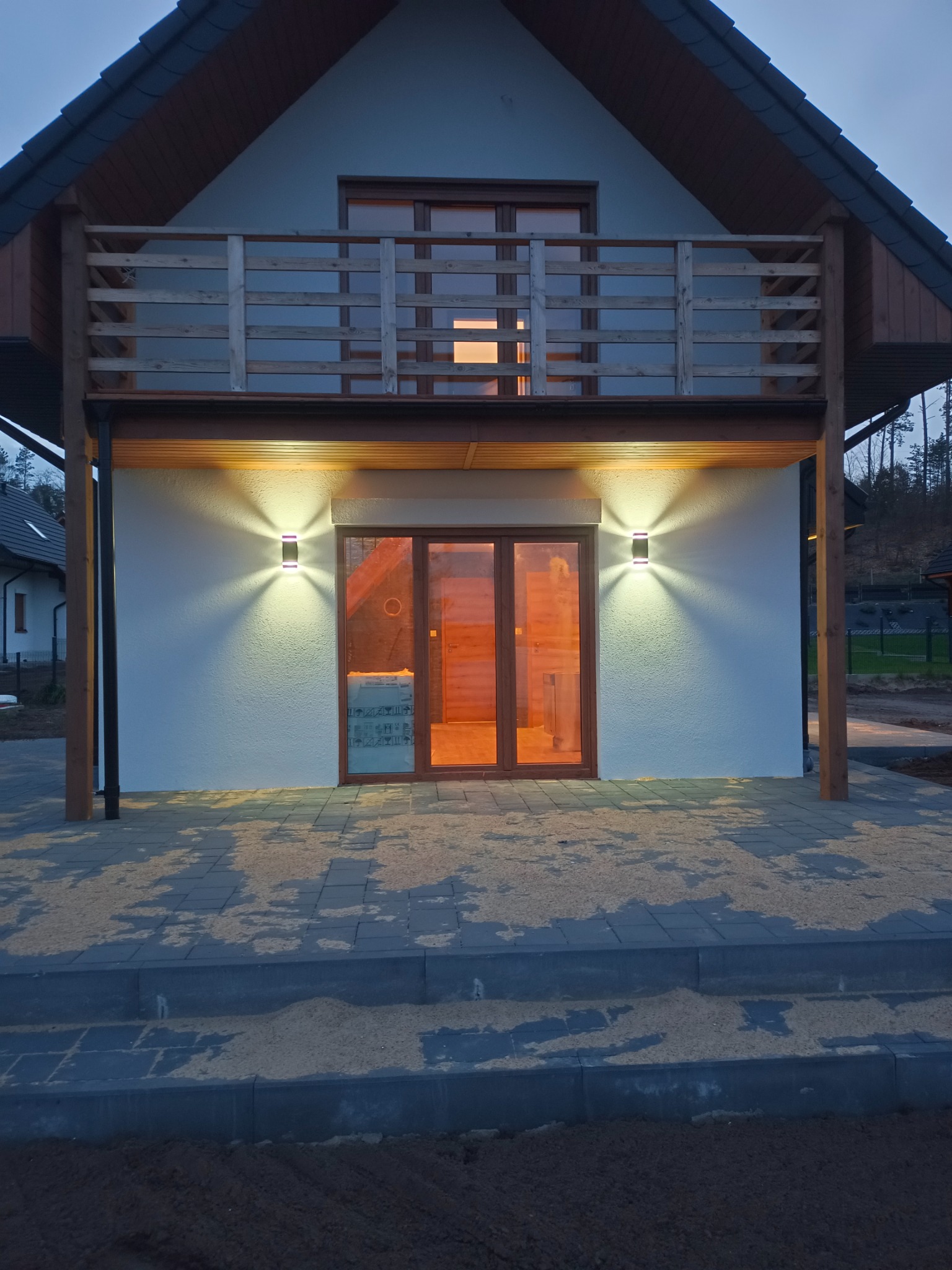 Nowoczesny dom jednorodzinny z balkonem i oświetleniem zewnętrznym, widoczne lampy elewacyjne po bokach drewnianych drzwi balkonowych, kostka brukowa na tarasie.