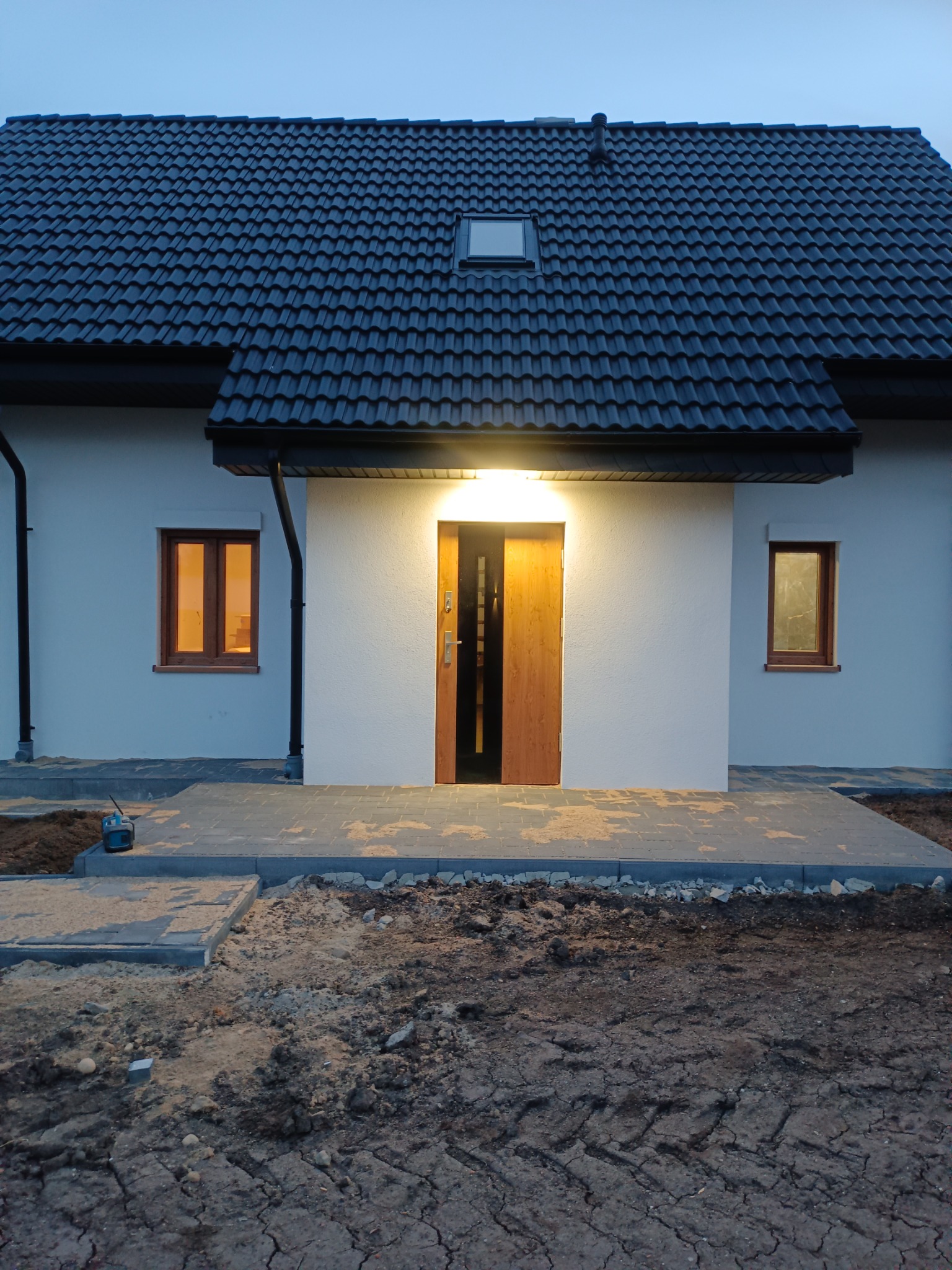 Nowoczesny dom jednorodzinny z ciemną dachówką i oświetlonym wejściem, widoczne prace brukarskie na podwórku, wieczorne oświetlenie w oknach.