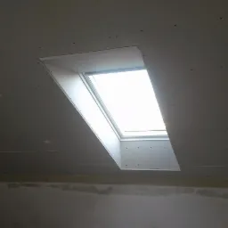 Montaż okna dachowego w suficie z płyt karton-gips, widoczne łączenia płyt. Jasne światło dzienne wpada przez okno.