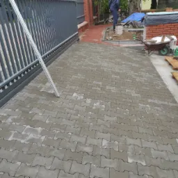 Ułożona szara kostka brukowa na podjeździe, widoczna brama przesuwna, w tle ceglany murek, taczka i konewki na paletach.