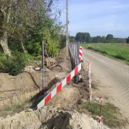 Budowa ogrodzenia panelowego z podmurówką: widoczne metalowe słupki, zbrojenie pod betonową podmurówkę, zabezpieczenia budowy w postaci taśm i barierek, obok droga z kostki.