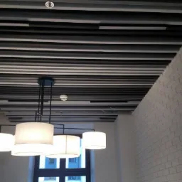 Nowoczesny sufit z listew w odcieniach szarości i bieli, oświetlony wiszącą lampą z białymi kloszami i widoczną instalacją pod sufitem, na tle białej ściany z cegły.