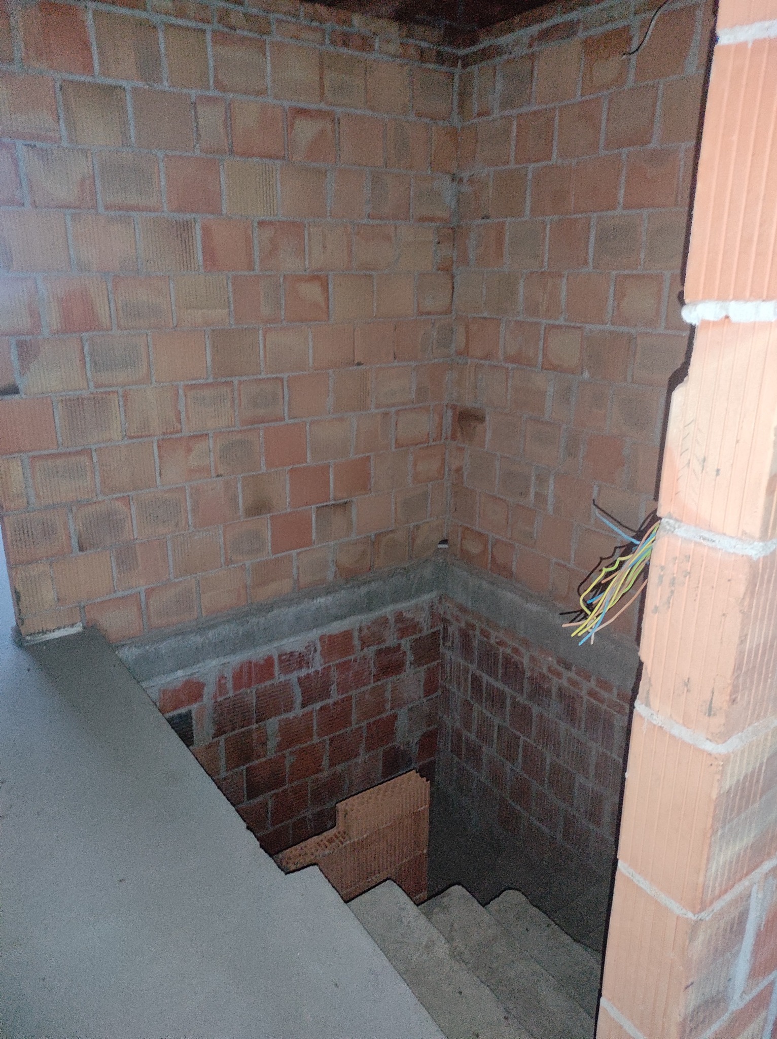 Murowana ściana z cegły ceramicznej w piwnicy, widoczne schody betonowe, wystające przewody elektryczne w kolorach żółtym, niebieskim i czarnym.