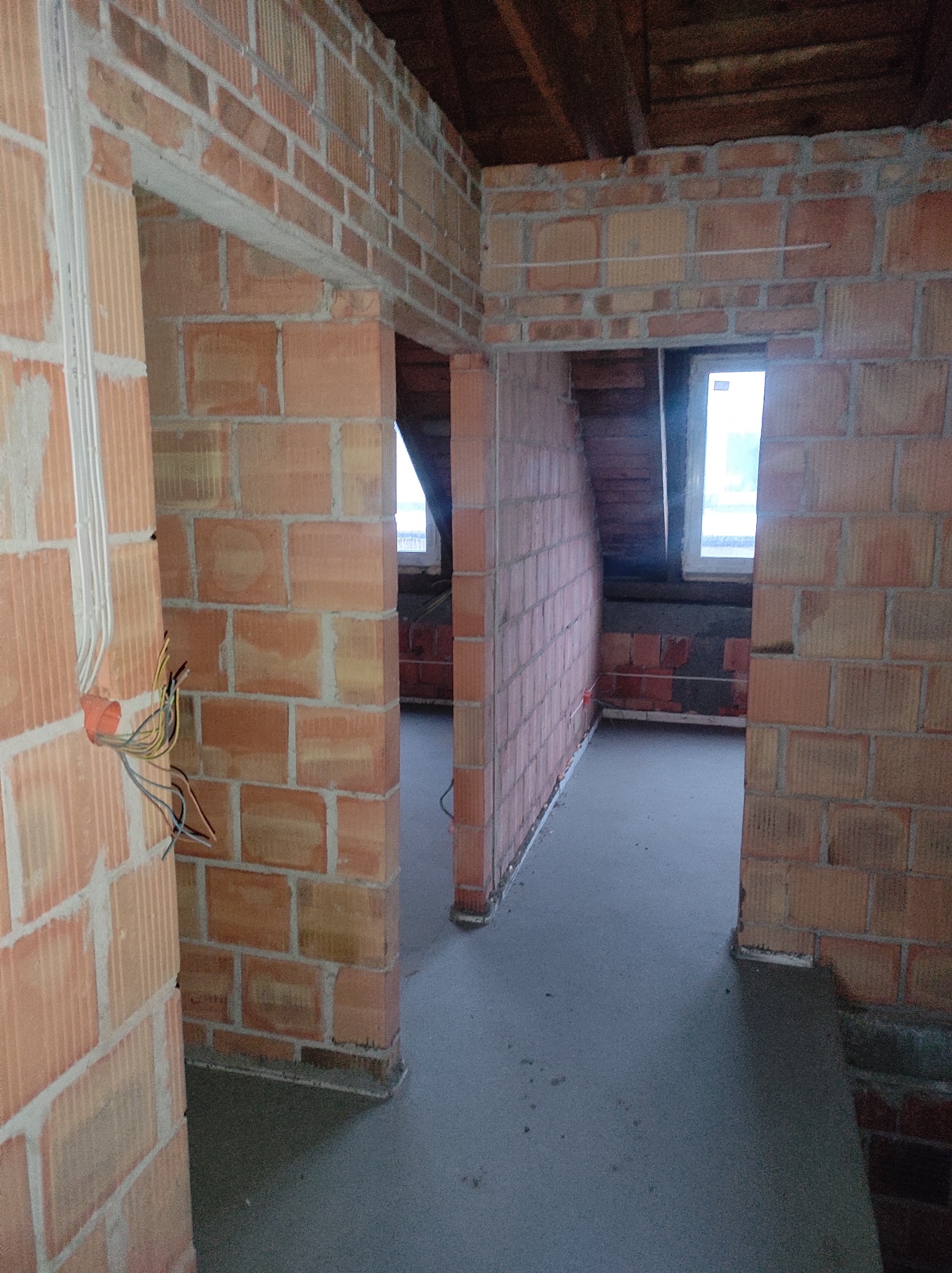 Wnętrze poddasza w trakcie budowy, ściany z czerwonej cegły, widoczne okno i instalacja elektryczna z przewodami.
