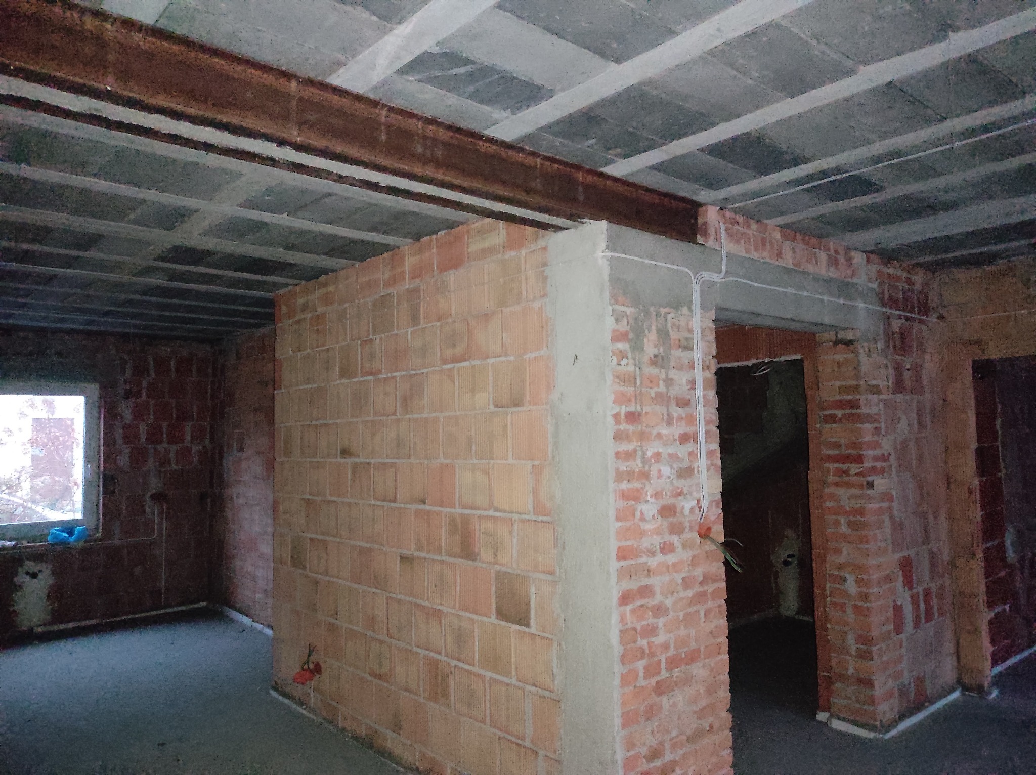Wnętrze w trakcie remontu: surowy strop z widoczną stalową belką, fragment ściany z czerwonej cegły z wyprowadzonymi przewodami elektrycznymi, surowa podłoga, okno w głębi pomieszczenia.