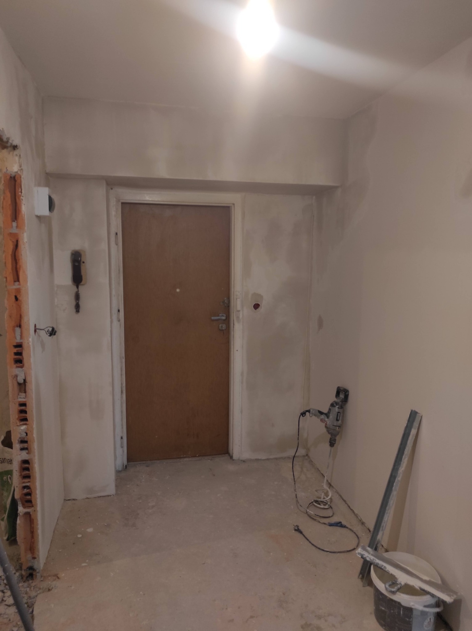 Wnętrze mieszkania w trakcie prac wykończeniowych: ściany świeżo pokryte gładzią, widoczna boczna ściana z odsłoniętą cegłą, narzędzia do nakładania gładzi oraz wiertarka przy ścianie.