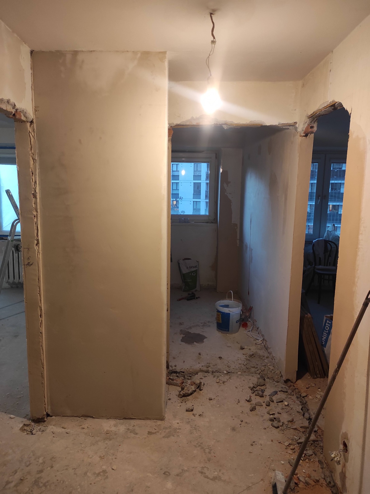 Wnętrze mieszkania w trakcie prac wykończeniowych: ściany pokryte świeżym tynkiem, widoczne ślady po kuciu, resztki gruzu na podłodze, odsłonięte przewody elektryczne, wiadro z farbą i worki...