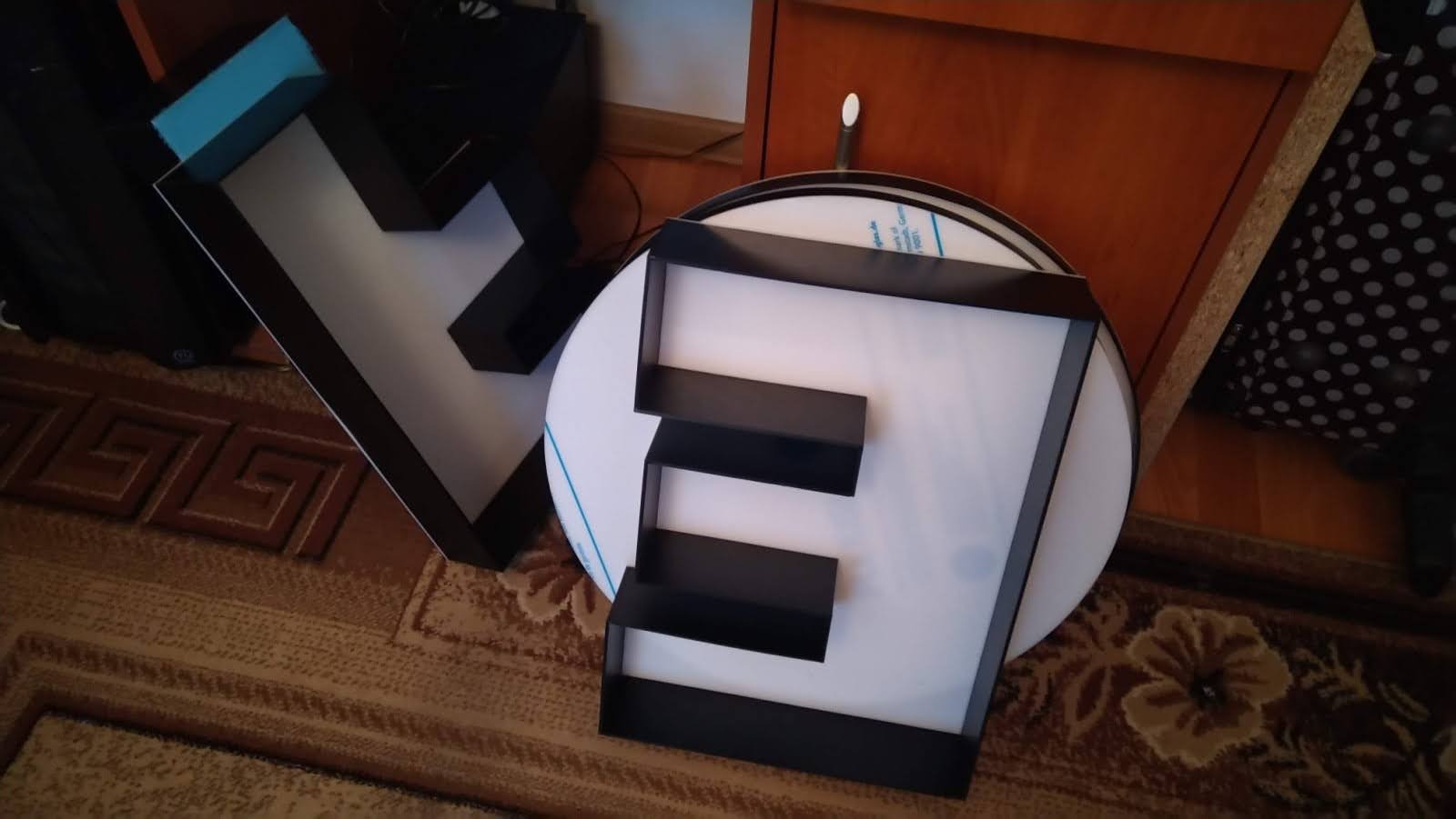 Litery przestrzenne 'V' i 'E' z czarnymi krawędziami i białym wnętrzem, leżące na dywanie i podłodze, częściowo oparte o meble.