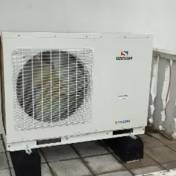 Zewnętrzna jednostka pompy ciepła monoblok Sinclair 10 kW, zamontowana na czarnych gumowych podstawach na szarej, kwadratowej kostce brukowej, z widocznym logo Sinclair i elementami instalacji...