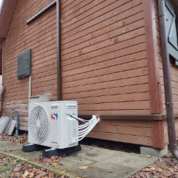 Zewnętrzna jednostka klimatyzacji Sinclair MultiSplit, zamontowana na betonowej płycie przed drewnianym domkiem letniskowym, widoczne liczne przewody łączące jednostkę z instalacją wewnątrz budynku...
