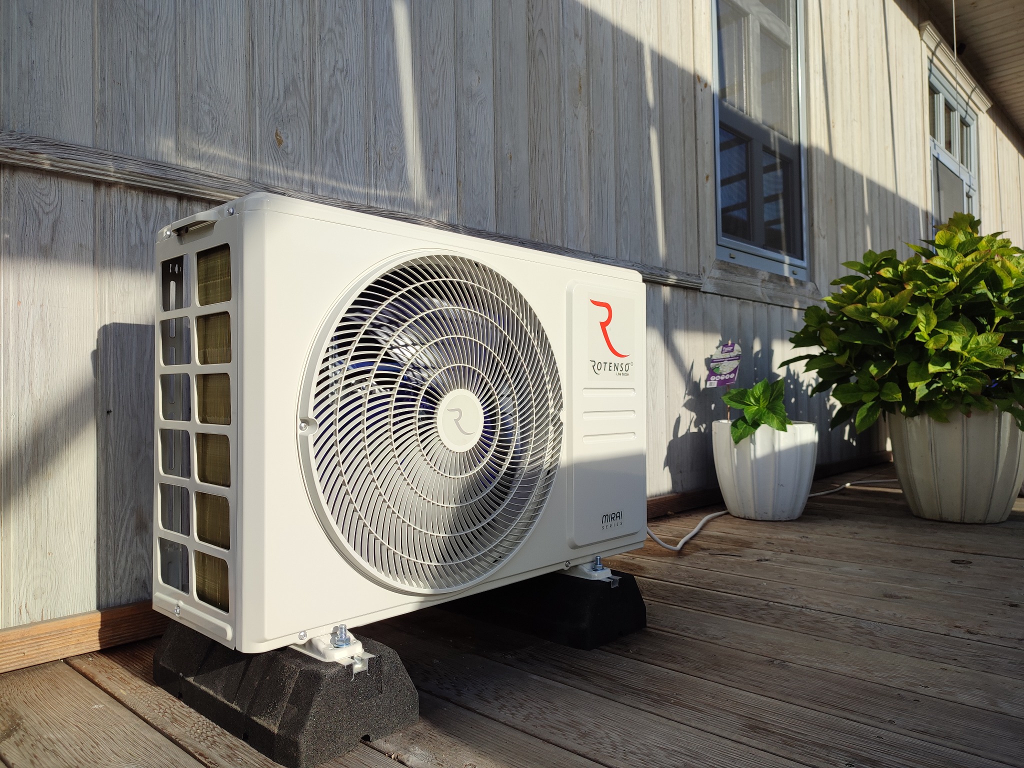 Zewnętrzna jednostka klimatyzacji Rotenso Mirai, zamontowana na czarnych podkładkach tłumiących drgania, na drewnianym tarasie przed domem z drewnianą elewacją i dwoma oknami. Obok jednostki...