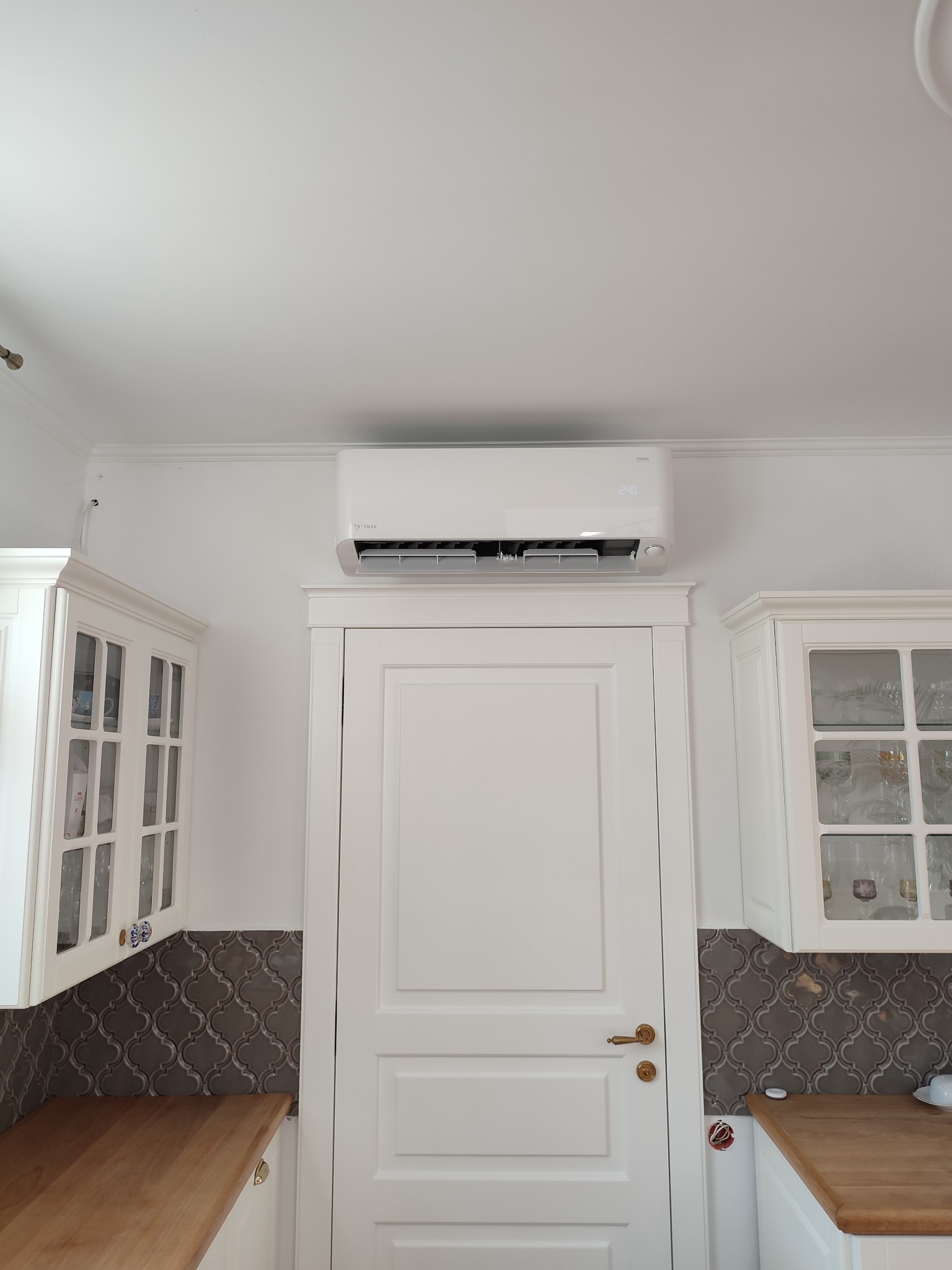 Biała jednostka wewnętrzna klimatyzacji Rotenso Mirai zamontowana nad drzwiami w kuchni z białymi szafkami i szarą mozaiką na ścianie. Na wyświetlaczu klimatyzatora widoczna temperatura 24 stopnie.