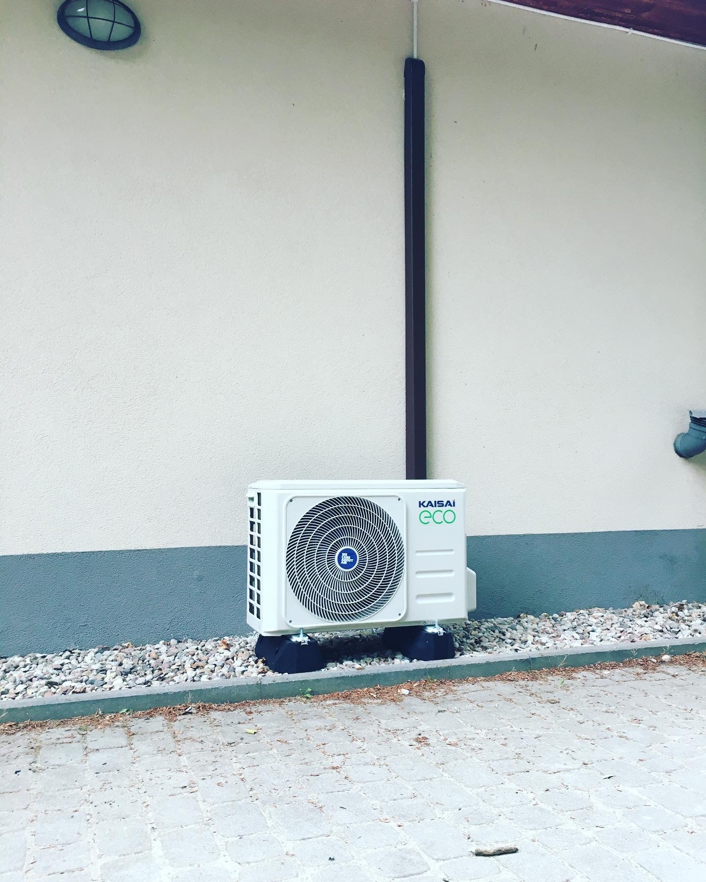 Biała jednostka zewnętrzna klimatyzacji Kaisai ECO 7 kW zamontowana na czarnych podkładkach, ustawiona na żwirze przy ścianie budynku z szarą lamperią.