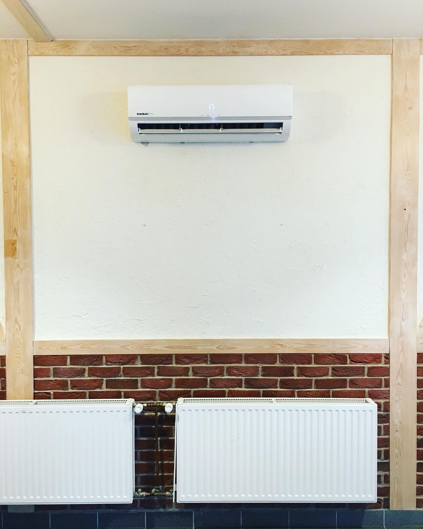 Biały klimatyzator Kaisai ECO zamontowany na kremowej ścianie, otoczony dekoracyjną ramą z jasnego drewna i pasem czerwonej cegły, widoczne dwa białe grzejniki panelowe.