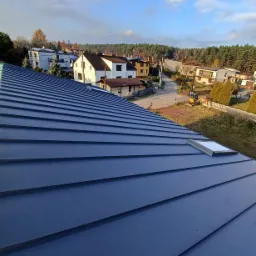 Nowy, grafitowy dach z blachy na tle zabudowy jednorodzinnej i błękitnego nieba z chmurami. Widoczny świetlik dachowy.