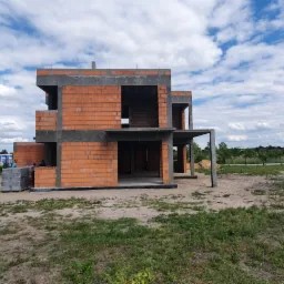 Surowy stan dwukondygnacyjnego budynku z czerwonej cegły, widoczny na tle pochmurnego nieba, z elementami betonowymi i paletą cegieł w pobliżu.