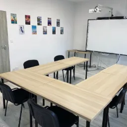 Sala lekcyjna z jasnym drewnianym stołem w kształcie podkowy, czarnymi krzesłami, interaktywną tablicą i projektorem, ozdobiona plakatami z motywami podróżniczymi.