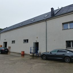 Stakochem - Duży, biały budynek magazynowy z kilkoma bramami wjazdowymi, zaparkowanymi samochodami i vanem na zewnątrz w pochmurny dzień.