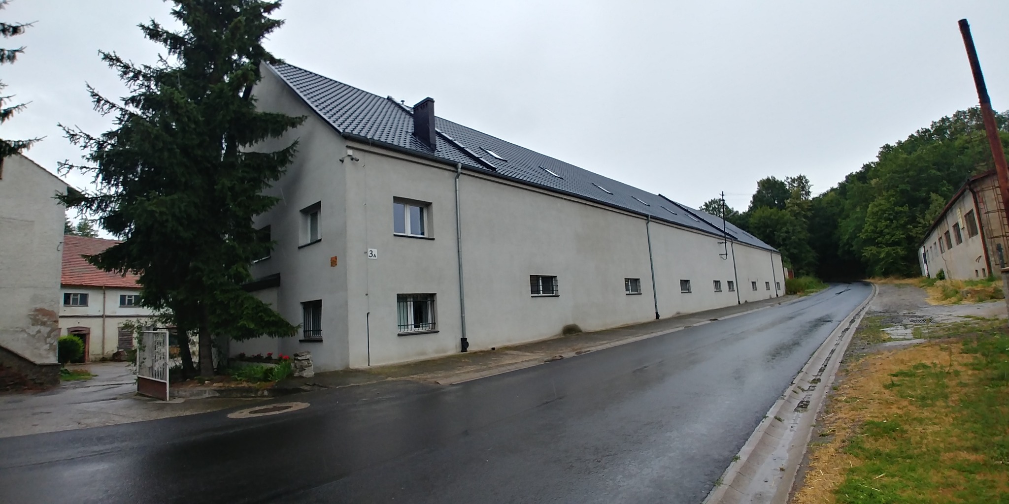 Długi, jednopiętrowy budynek magazynowy z szarą elewacją i czarnym dachem, widoczny z perspektywy ulicy w pochmurny dzień.