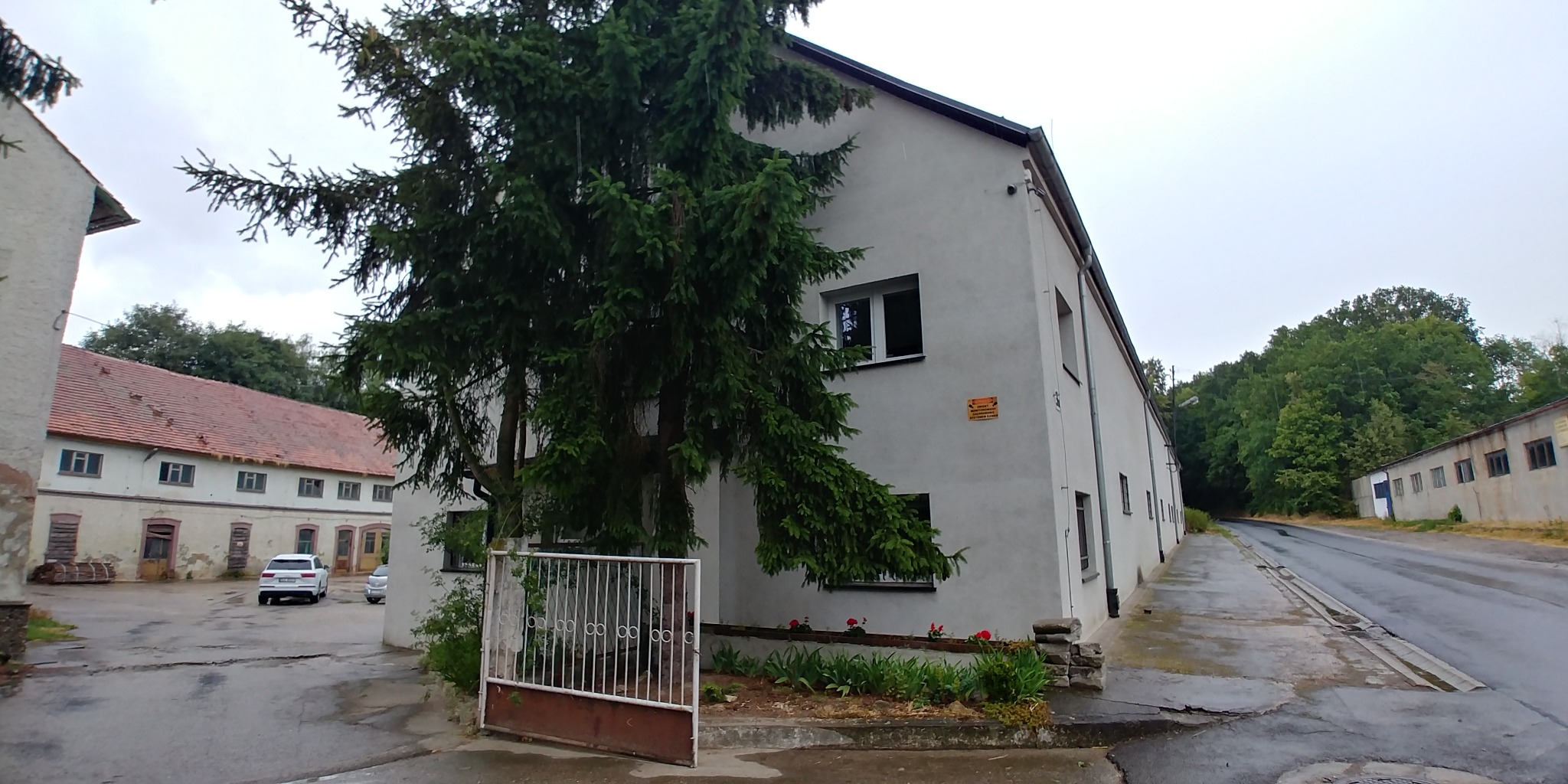 Zewnętrzny widok magazynu z szarą elewacją, widoczne drzewo iglaste i ogrodzenie, w tle budynek z czerwoną dachówką i zaparkowane samochody.