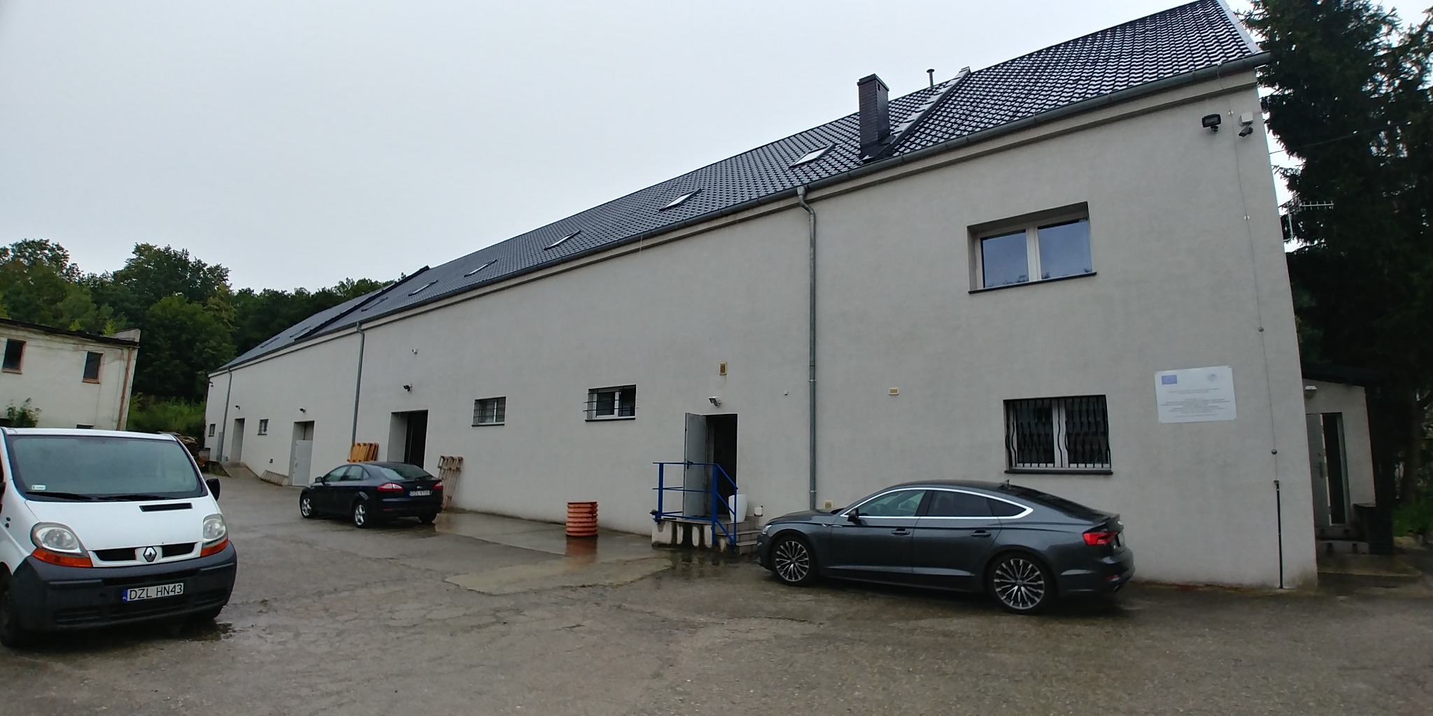 Duży, biały budynek magazynowy z kilkoma bramami wjazdowymi, zaparkowanymi samochodami i vanem na zewnątrz w pochmurny dzień.