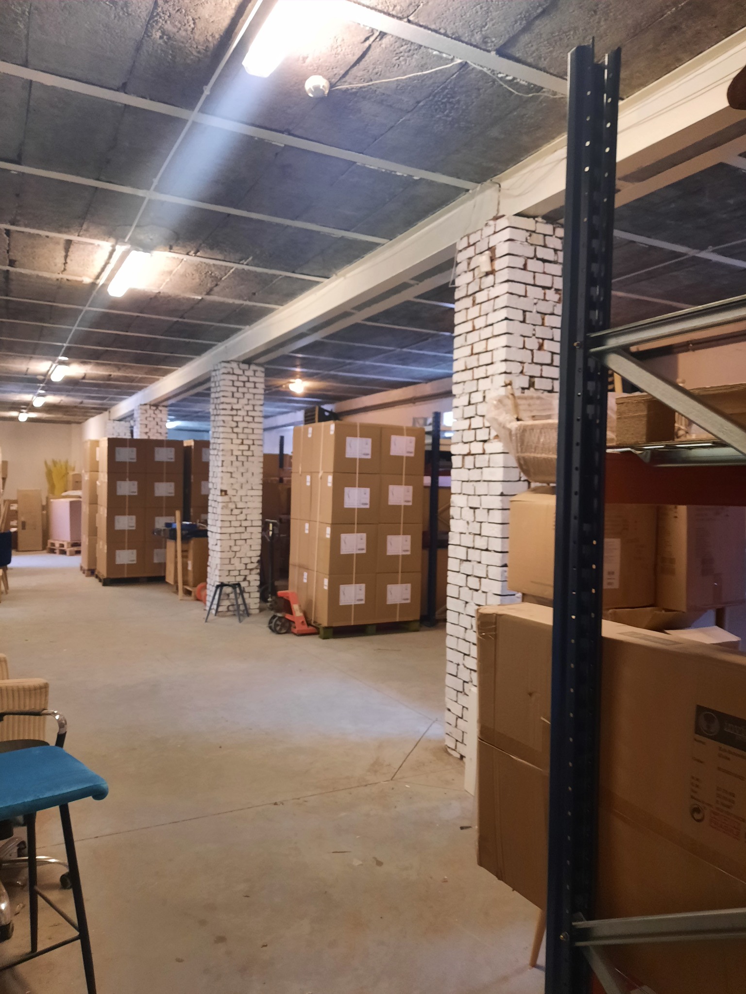 Wnętrze magazynu z rzędami zapakowanych kartonów na paletach, oświetlone lampami sufitowymi, widoczne regały i kolumny z białej cegły.