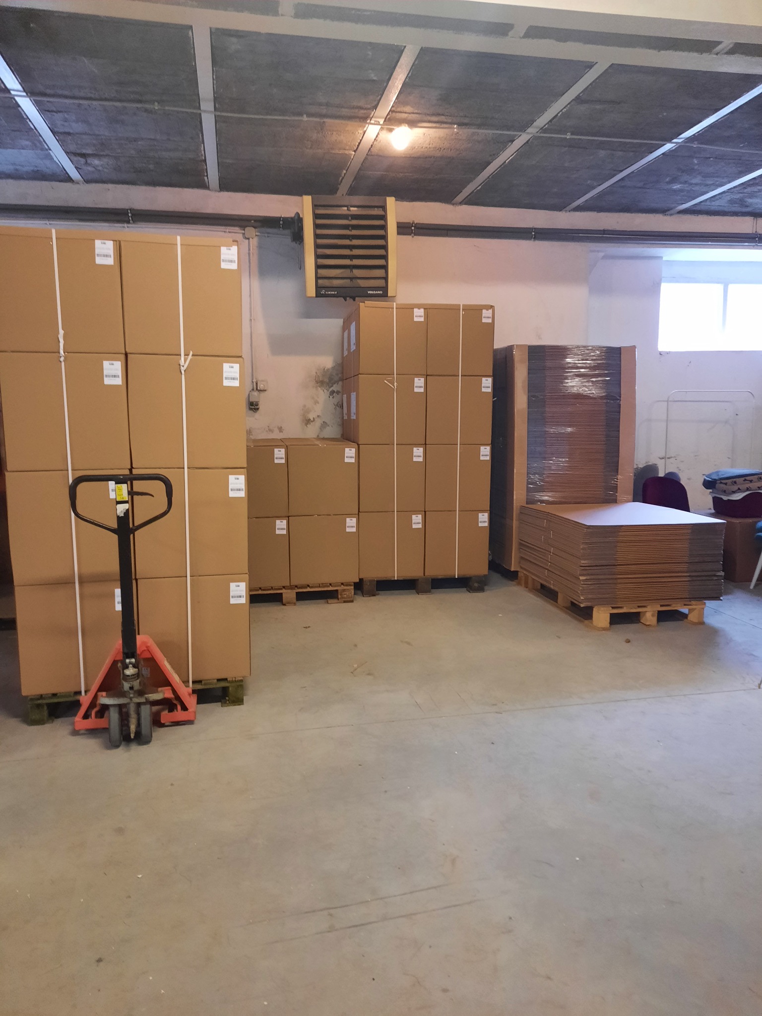 Wnętrze magazynu z paletami wypełnionymi kartonami, część z nich zabezpieczona folią stretch, wózek paletowy z pomarańczowym podnośnikiem.