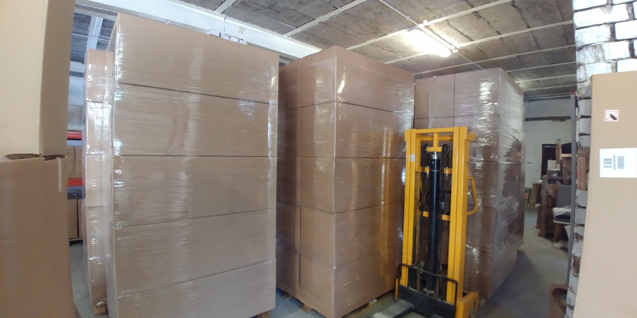 Spiętrzone, owinięte folią palety z kartonami w magazynie, obok wózek widłowy, surowy betonowy sufit.