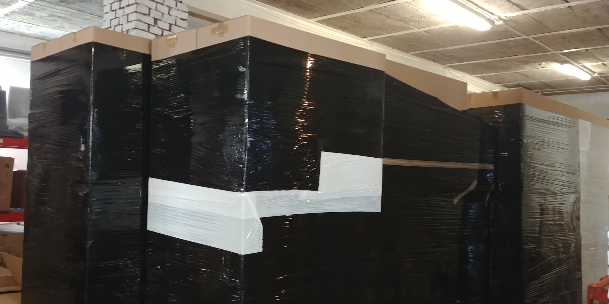 Wysokie, czarne paczki owinięte folią stretch i zabezpieczone białą taśmą, ustawione w rzędzie w magazynie z widocznym betonowym sufitem i fragmentem ceglanej ściany.