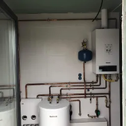 Nowoczesna kotłownia z piecem gazowym Buderus, zasobnikiem ciepłej wody i rozbudowaną instalacją miedzianą, widoczna w pomieszczeniu z białymi płytkami i elementami w stanie surowym.