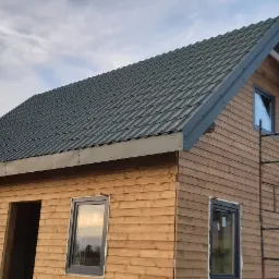 Dom w trakcie budowy z drewnianą elewacją i nowym, grafitowym dachem z blachy trapezowej; widoczne rusztowanie i odbicie zachodzącego słońca w oknach.