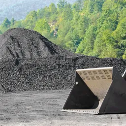 Stos czarnego węgla na tle zielonych drzew i niebieskich gór, z łyżką ładowarki na pierwszym planie.